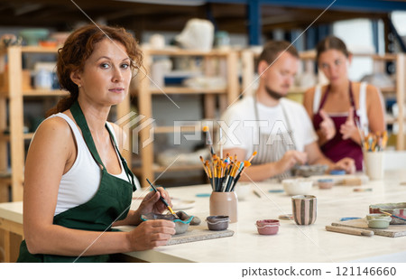 Woman learns to make designs on clay plate using brush and paint in pottery workshop 121146660