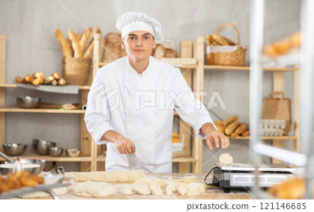 Young male baker cutting and weighing dough on scales in bakehouse 121146685