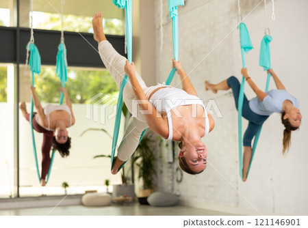Fit and beautiful young woman hanging upside down while practicing aerial yoga during group class in fitness club 121146901