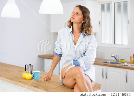 Lovely sexy girl in shirt posing while sitting on the table of modern kitchen 121146923