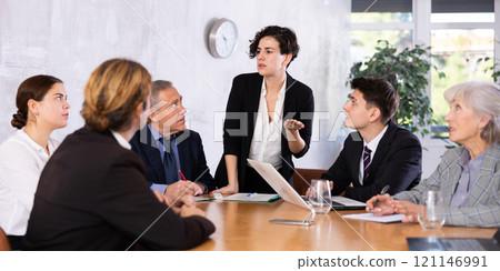 Concentrated young adult old office workers sitting around the table and looking at screen while Latin female using projector remote control in office 121146991