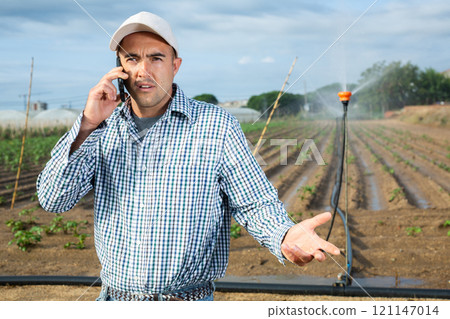 Focused farmer talking on phone while standing on farm field Focused farmer talking on phone while standing on farm field 121147014