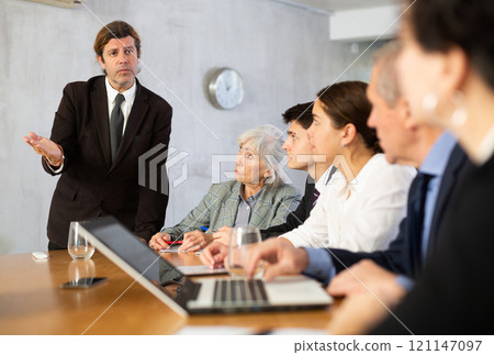 Dissatisfied male boss telling off office workers of different ages using notebooks at table in office 121147097