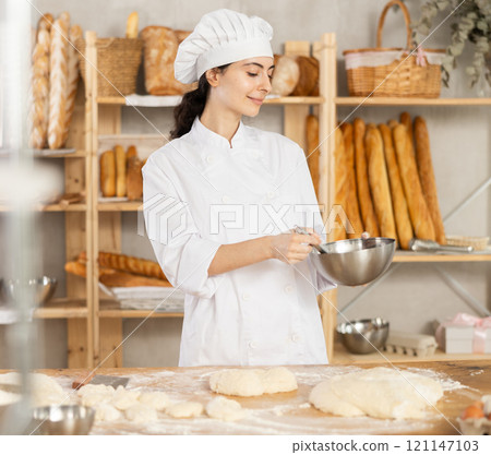 Baker girl in white uniform and chefs hat with bowl and whisk in her hands 121147103