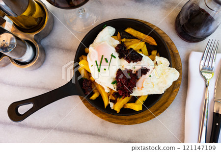 There is portion of French fries, fried eggs and piece of boutifara in pan. 121147109
