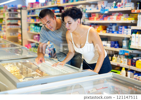 Positive couple choosing frozen food in refrigerated display cases in supermarket 121147511