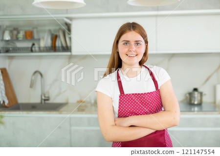 Portrait of long-haired young woman in red apron with polka dots, standing in kitchen Portrait of long-haired young woman in red apron with polka dots, standing in kitchen 121147554