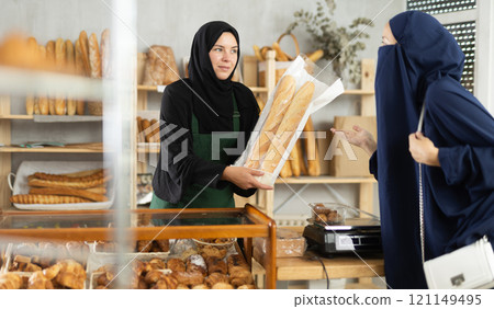 Selling fresh bread in an oriental bakery - girl seller hands bag of baguettes to female customer 121149495