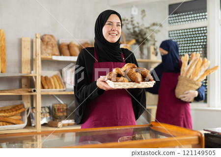 Young Muslim saleswoman demonstrates croissants while another woman stocks baguettes 121149505