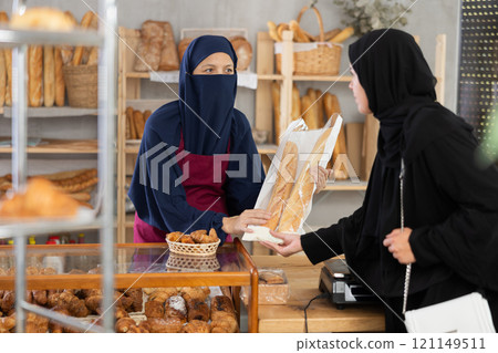 Niqab-wearing bakery worker handing baguettes to female customer Niqab-wearing bakery worker handing baguettes to female customer 121149511