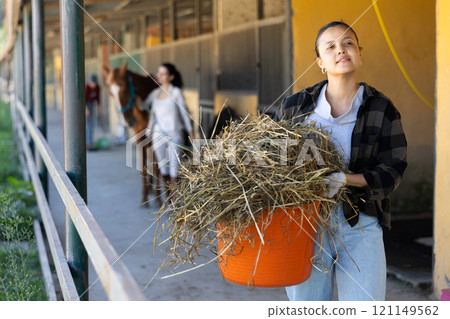 Asian young female worker walks go through corridor of stable, carrying basket with hay Asian young female worker walks go through corridor of stable, carrying basket with hay 121149562