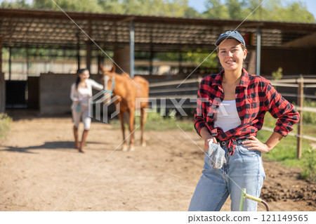 Positive female stable worker stands near wooden fence of paddock and watches horse. Positive female stable worker stands near wooden fence of paddock and watches horse. 121149565