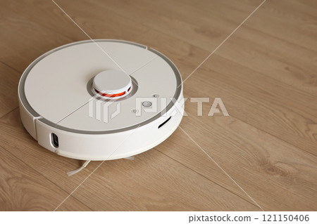 Top view of white wireless autonomous robotic vacuum cleaner on wooden floor. Smart cleaning technology. Self-propelled cleaning robot. Floor cleaning system. Household remote control appliances. Top view of white wireless autonomous robotic vacuum cleaner on wooden floor. Smart cleaning technology. Self-propelled cleaning robot. Floor cleaning system. Household remote control appliances. 121150406