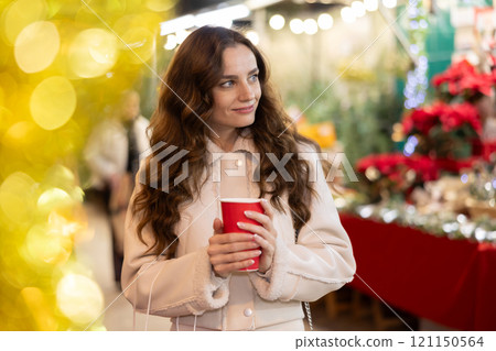 Cheerful woman with paper cup of hot tea walking at Christmas street fair Cheerful woman with paper cup of hot tea walking at Christmas street fair 121150564