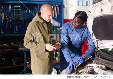 Worried client talking with specialist about repairing his car engine at workshop 121150685