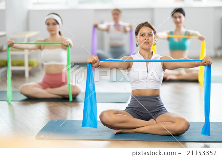 Young woman bring together and spread straightened arms at chest level with wide resistance band 121153352
