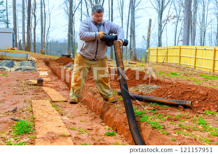 Man is working on installing drainage system in backyard, in freshly dug trench to enhance water flow management. 121157951