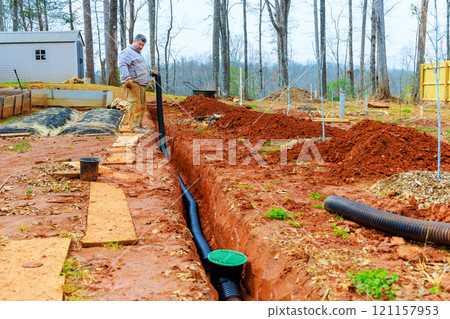 Technical worker works on drainage installation in his garden, managing soil, pipes amidst during construction Technical worker works on drainage installation in his garden, managing soil, pipes amidst during construction 121157953