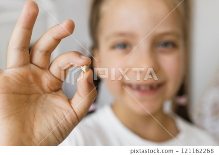 Smiling little Caucasian girl proudly holding her lost baby tooth at home, showcasing a milestone. The focus is on her delight and the symbol of growing up. 121162268