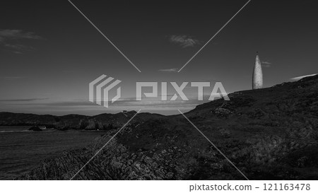 A white, tall, slender marker stands on a grassy cliff. Dark, rugged rocks form the cliff face. Sea stretches to a distant coastline. 121163478