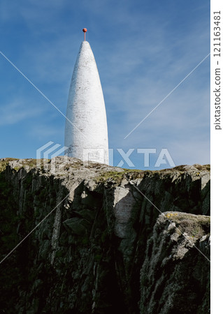 A conical, white navigation marker is positioned on a rugged, dark clifftop. The day is sunny and the sky is mostly clear. A conical, white navigation marker is positioned on a rugged, dark clifftop. The day is sunny and the sky is mostly clear. 121163481