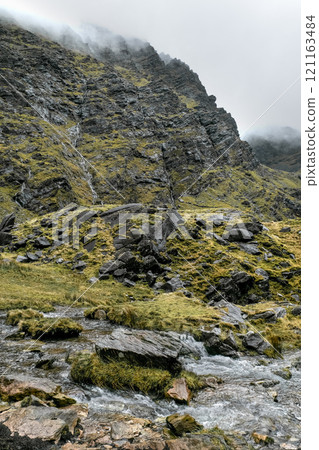 A rugged hillside rises steeply with patches of grass and rocks, while a gentle stream flows over stones. Mist shrouds the mountaintop, creating a tranquil atmosphere. A rugged hillside rises steeply with patches of grass and rocks, while a gentle stream flows over stones. Mist shrouds the mountaintop, creating a tranquil atmosphere. 121163484