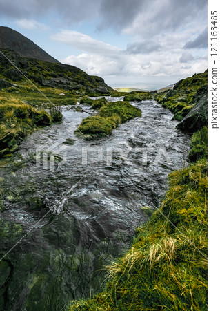 Clear water runs swiftly over rocks. The stream is nestled between grassy, rocky hills. Clear water runs swiftly over rocks. The stream is nestled between grassy, rocky hills. 121163485