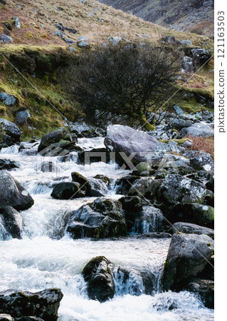 Water cascades over numerous rocks in a riverbed. The surrounding area features low-lying vegetation and distant rolling hills. A single tree is visible. Water cascades over numerous rocks in a riverbed. The surrounding area features low-lying vegetation and distant rolling hills. A single tree is visible. 121163503