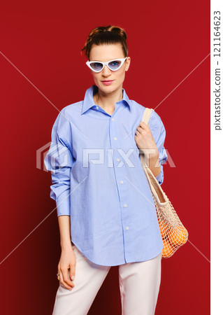 Fashionable woman posing in blue oversized shirt against a vibrant red background 121164623