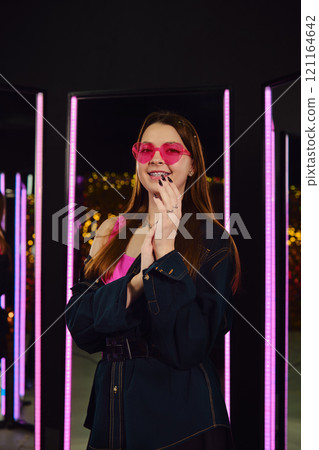 Young woman poses confidently in a trendy outfit with vibrant lights in the background Young woman poses confidently in a trendy outfit with vibrant lights in the background 121164642