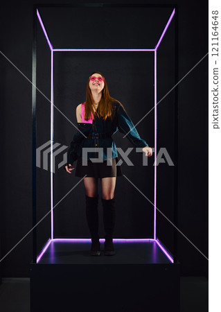 Young woman poses in modern lighted box with neon accents, expressing confidence and style Young woman poses in modern lighted box with neon accents, expressing confidence and style 121164648