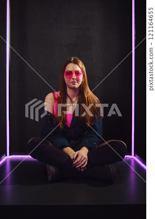 Young woman poses stylishly in neon-lit studio wearing pink sunglasses 121164655