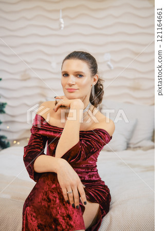 Elegant woman in a red velvet dress poses thoughtfully in a cozy interior setting 121164661