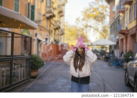 A Cheerful Young Woman Dressed in Stylish Winter Attire is Posing on a Bustling City Street 121164714