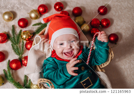 Merry Christmas and Happy New Year. A cheerful little child in a New Year's costume lies in a basket with Christmas tree balls Merry Christmas and Happy New Year. A cheerful little child in a New Year's costume lies in a basket with Christmas tree balls 121165963