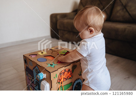 Happy toddler baby boy is playing with a busyboard cube on the nursery floor. A child aged one year 121165980