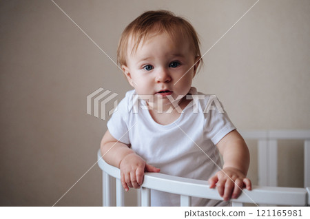Beautiful little girl in a crib in a cozy room in learning to stand with support. Cute girl standing in a crib at home 121165981
