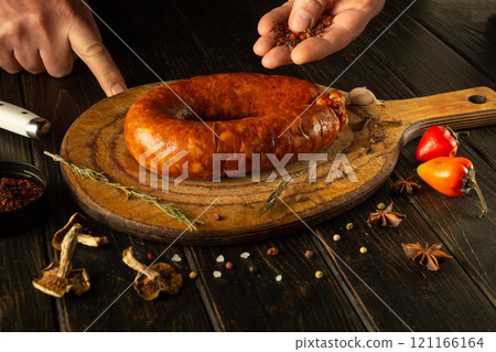 Skilled hands prepare a flavorful sausage on a wooden board, garnished with fresh herbs and spices, surrounded by colorful peppers, creating an inviting culinary atmosphere 121166164