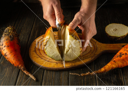 Two hands expertly cut a vibrant orange beet vegetable on a wooden board surrounded by other freshly harvested produce. The warm kitchen ambiance highlights the rich textures 121166165