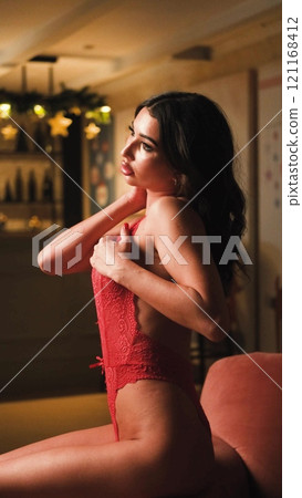 Beautiful brunette posing sexy on camera in red lace bodysuit while sitting on sofa Beautiful brunette posing sexy on camera in red lace bodysuit while sitting on sofa 121168412