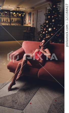 Beautiful brunette posing sexy on camera in red lace bodysuit while sitting on sofa 121168427