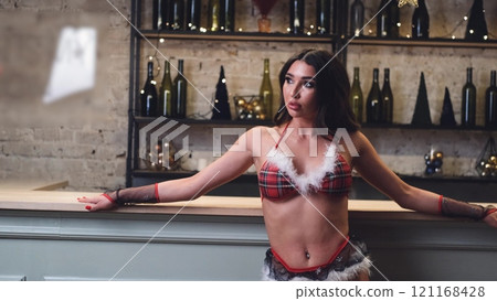 Sexy curly brunette posing in Santa Claus style lingerie, standing near the bar counter in a festively decorated room. Merry Christmas and Happy New Year Christmas holiday. 121168428
