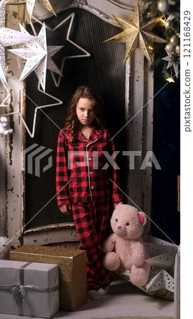 Portrait of sad dissatisfied little girl in night pajamas holding teddy bear in hands. Child disappointed with gifts from Santa Claus. Christmas night, festive atmosphere Portrait of sad dissatisfied little girl in night pajamas holding teddy bear in hands. Child disappointed with gifts from Santa Claus. Christmas night, festive atmosphere 121168429
