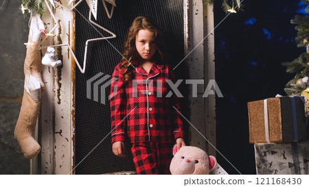 Portrait of sad dissatisfied little girl in night pajamas holding teddy bear in hands. Child disappointed with gifts from Santa Claus. Christmas night, festive atmosphere 121168430