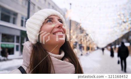 Portrait of a beautiful young woman in winter clothes. The woman smiles happily against the background of a snowy street decorated with Christmas garlands. 121168452