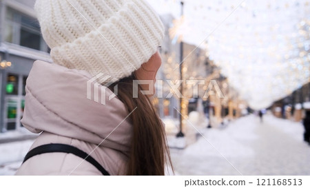 Portrait of a beautiful young woman in winter clothes. The woman smiles happily against the background of a snowy street decorated with Christmas garlands. Portrait of a beautiful young woman in winter clothes. The woman smiles happily against the background of a snowy street decorated with Christmas garlands. 121168513