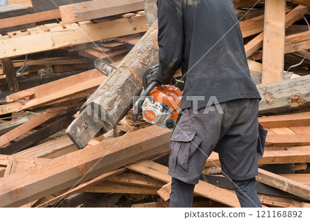 A worker cutting wood from a demolished old wooden house with an electric saw 121168892