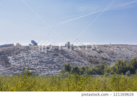 A large landfill site with garbage trucks and birds flying overhead. The image depicts the environmental impact of human waste and the need for sustainable solutions. 121169626