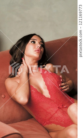 Beautiful brunette posing sexy on camera in red lace bodysuit while sitting on sofa Beautiful brunette posing sexy on camera in red lace bodysuit while sitting on sofa 121169773