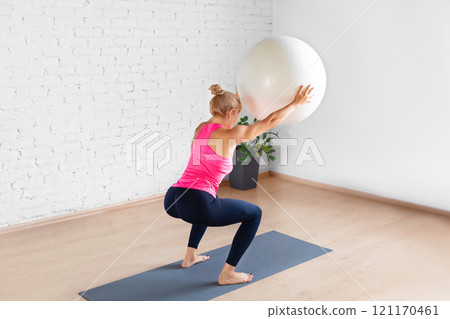 Fit Caucasian woman in pink tank top practice squat exercise using fitness ball in loft studio indoor, rear view. 121170461